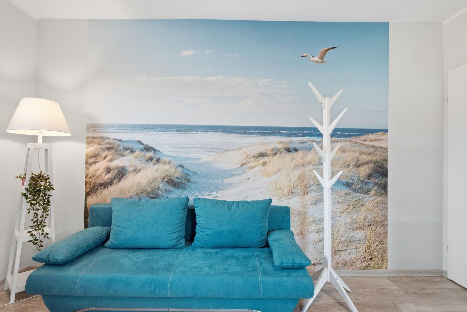 Ein blauer Sofa mit Kissen vor einer Wandtapete mit Strandmotiv und einem weißen Kleiderständer.