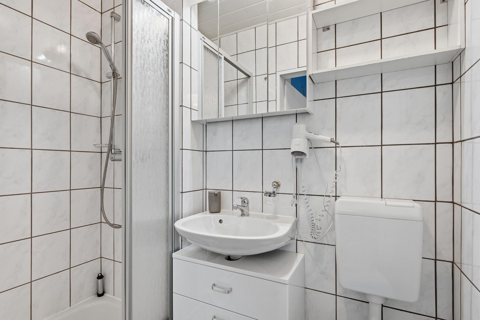 Badezimmer mit Dusche, Waschbecken und Toilette. Weiße Fliesen und Spiegelschrank.