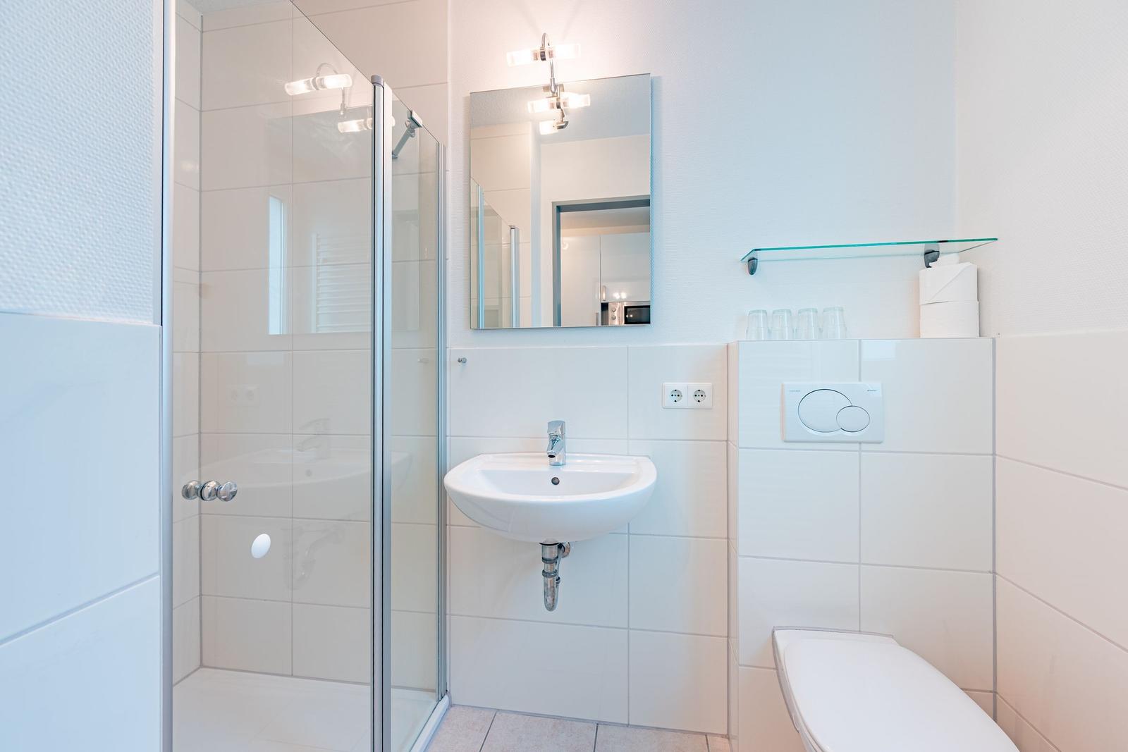 Badezimmer mit Dusche, Waschbecken und Toilette. Weiße Fliesen und Spiegel.
