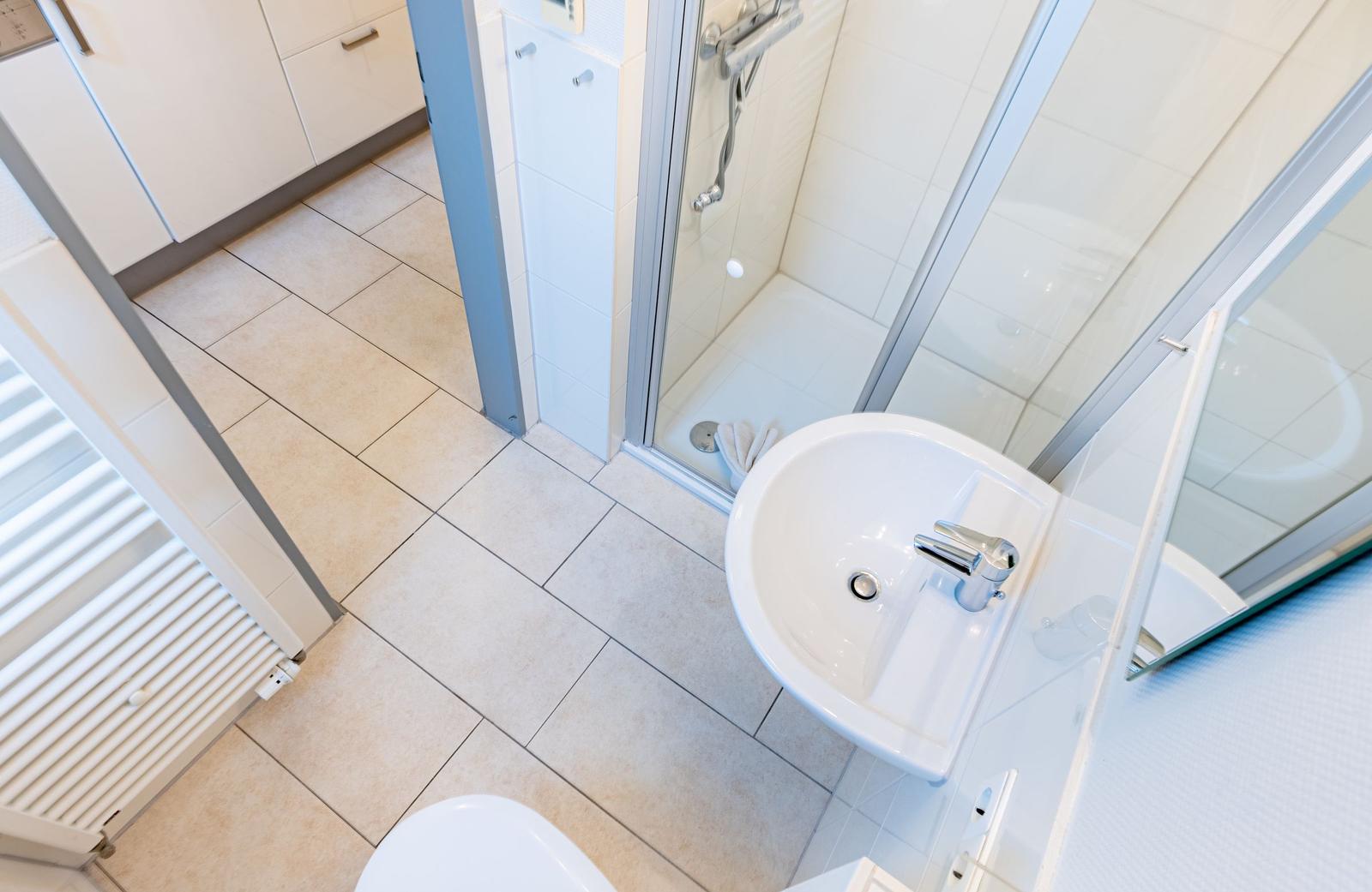 Badezimmer mit Dusche, Waschbecken und Toilette. Fliesenboden und weiße Einrichtung.