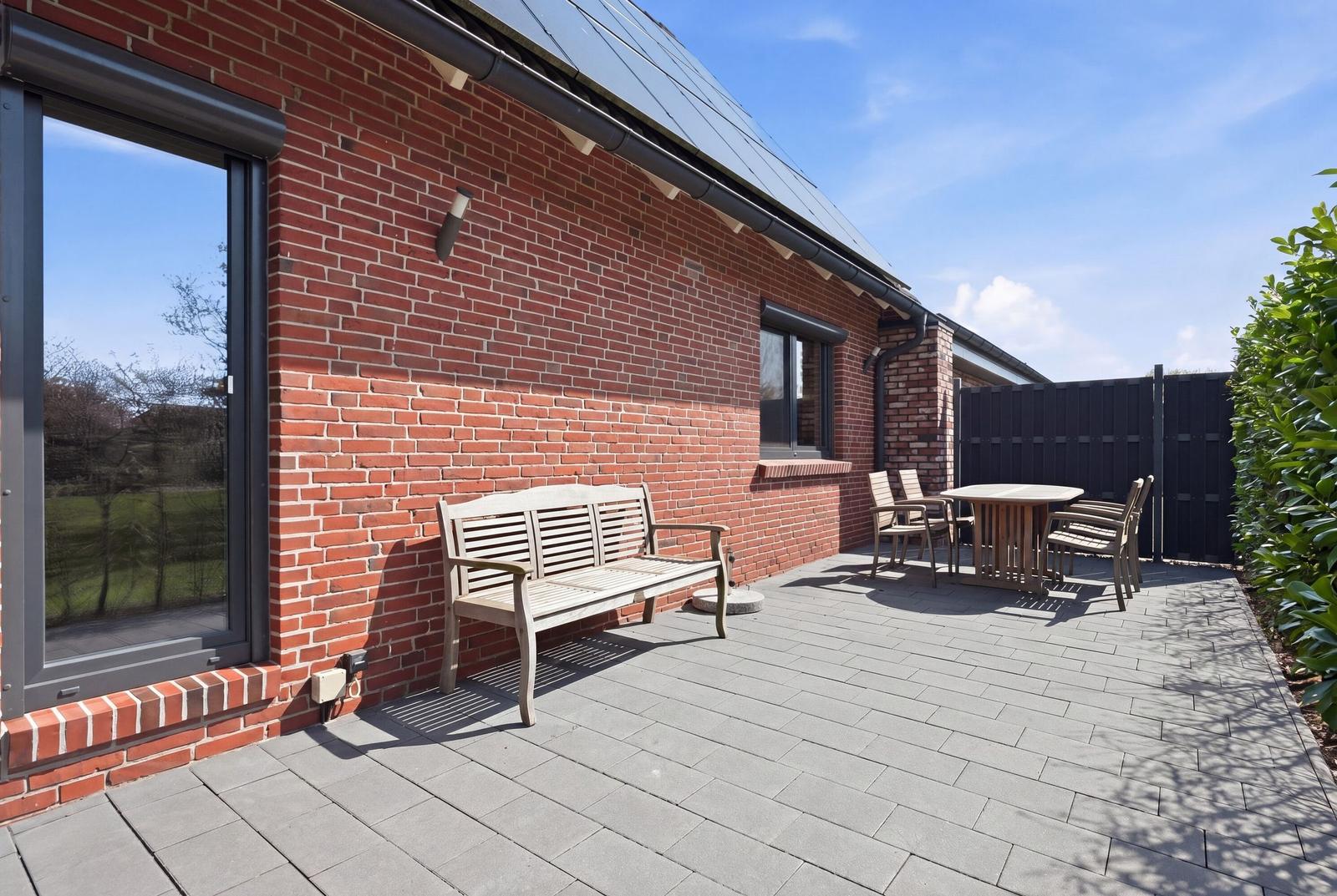 Terrasse mit Holzbank, Tisch und Stühlen neben rotem Backsteinhaus unter blauem Himmel.