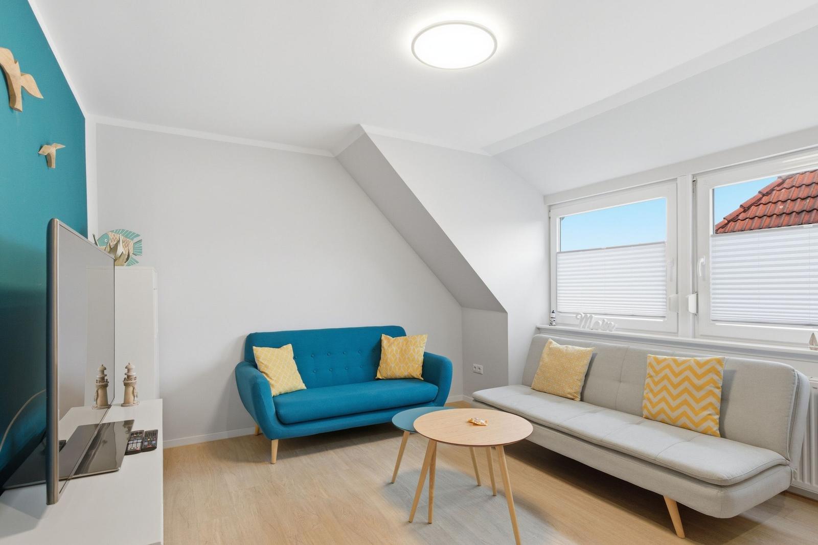 Wohnzimmer mit blauem Sofa, grauem Sofa und Holztisch. Fenster mit Jalousien und TV.