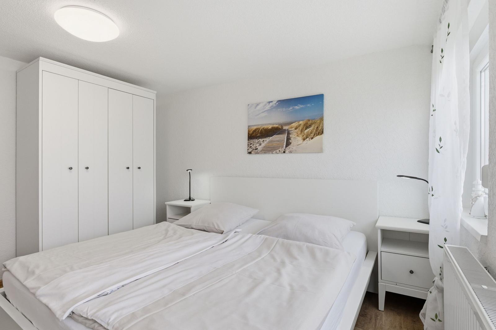 Schlafzimmer mit weißem Bett, Kleiderschrank und Wandbild. Zwei Nachttische mit Lampen.