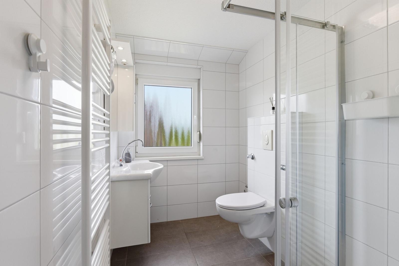 Badezimmer mit Dusche, Toilette und Waschbecken. Fenster mit getöntem Glas.