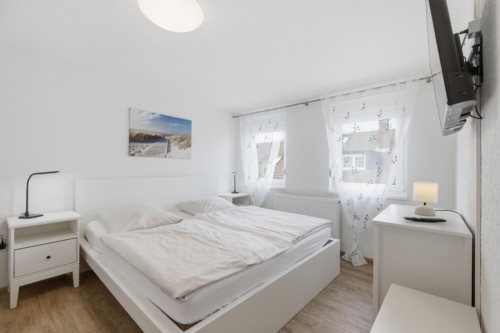 Schlafzimmer mit weißem Bett, Nachttischen und Fenster mit weißen Vorhängen.