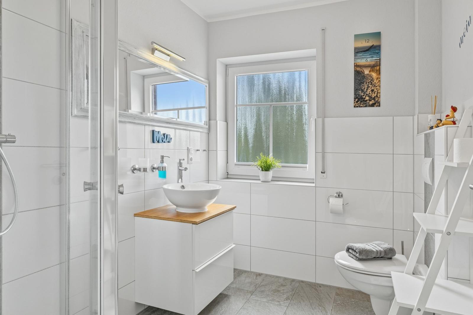 Badezimmer mit Dusche, Waschtisch und Toilette. Fenster mit Pflanze und Wandbild.