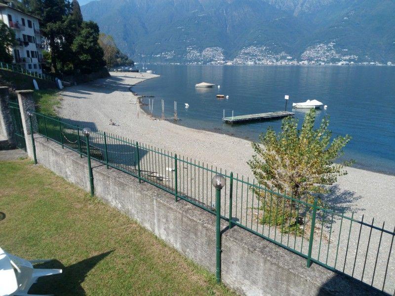 Blick auf einen Strand mit Steg und Booten am See. Grüner Zaun und Bäume im Vordergrund.
