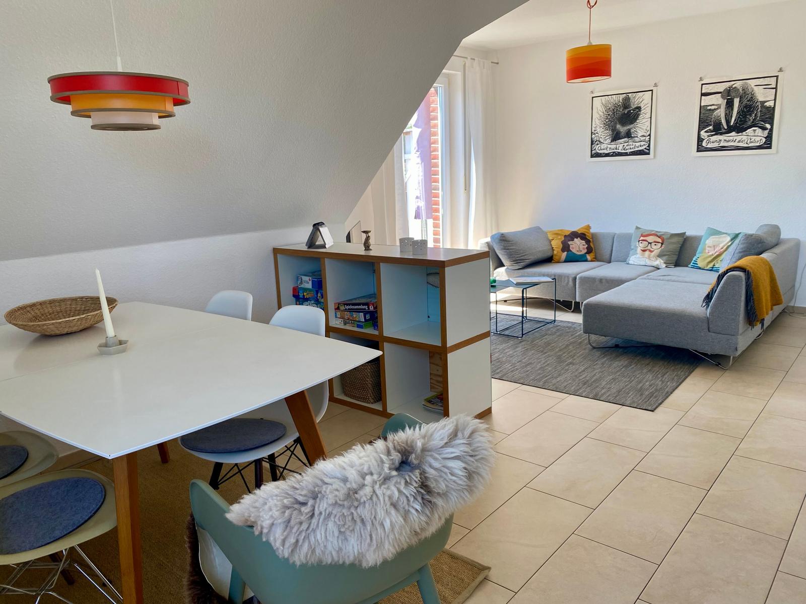 Wohn- und Essbereich mit Sofa, Tisch und Stühlen. Deckenleuchte und Wandbilder sichtbar.