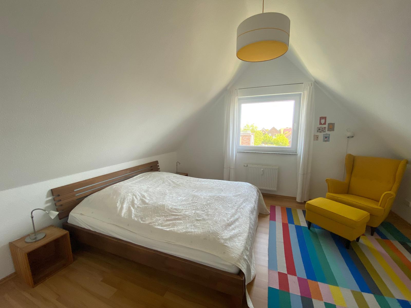 Schlafzimmer mit Dachschrägen, Bett, gelber Sessel und buntem Teppich.
