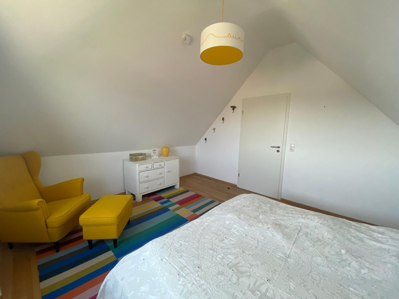 Schlafzimmer mit gelber Sitzgelegenheit, buntem Teppich und weißem Schrank unter schräger Decke.