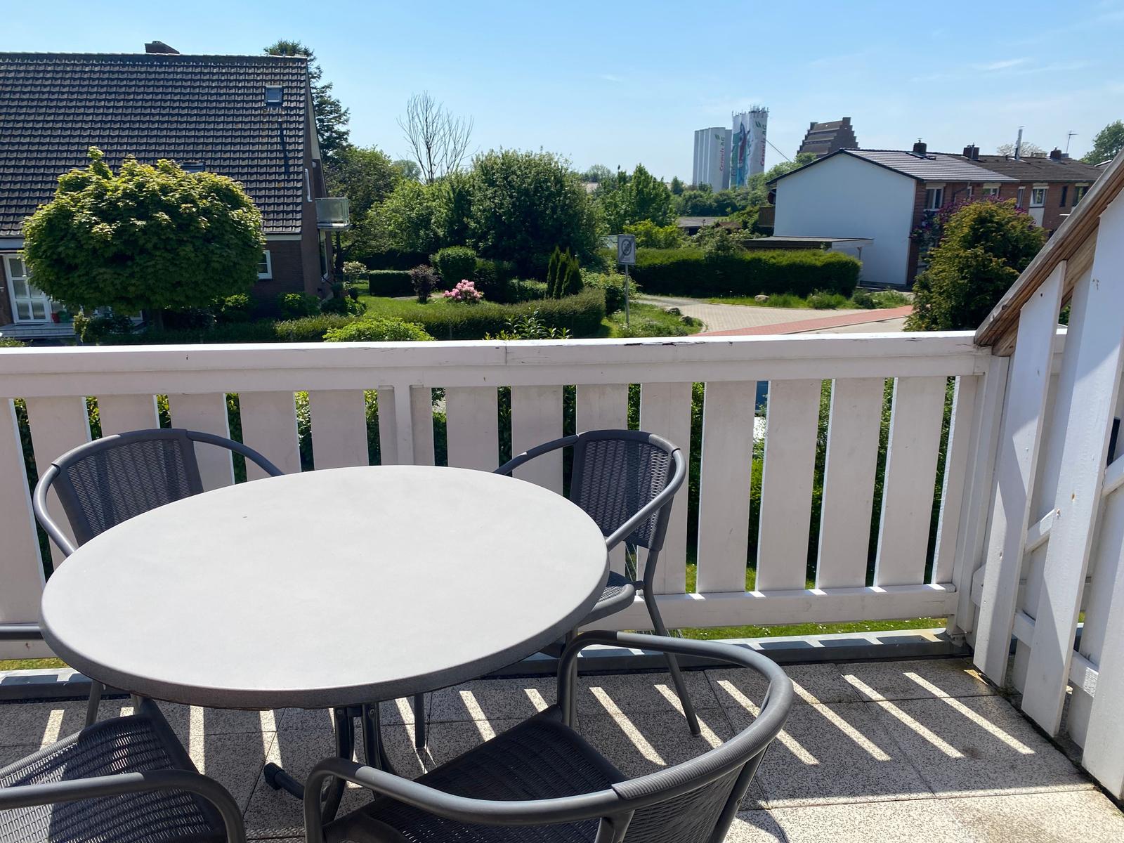 Terrasse mit runden Tisch und drei Stühlen. Blick auf grüne Gärten und Häuser.