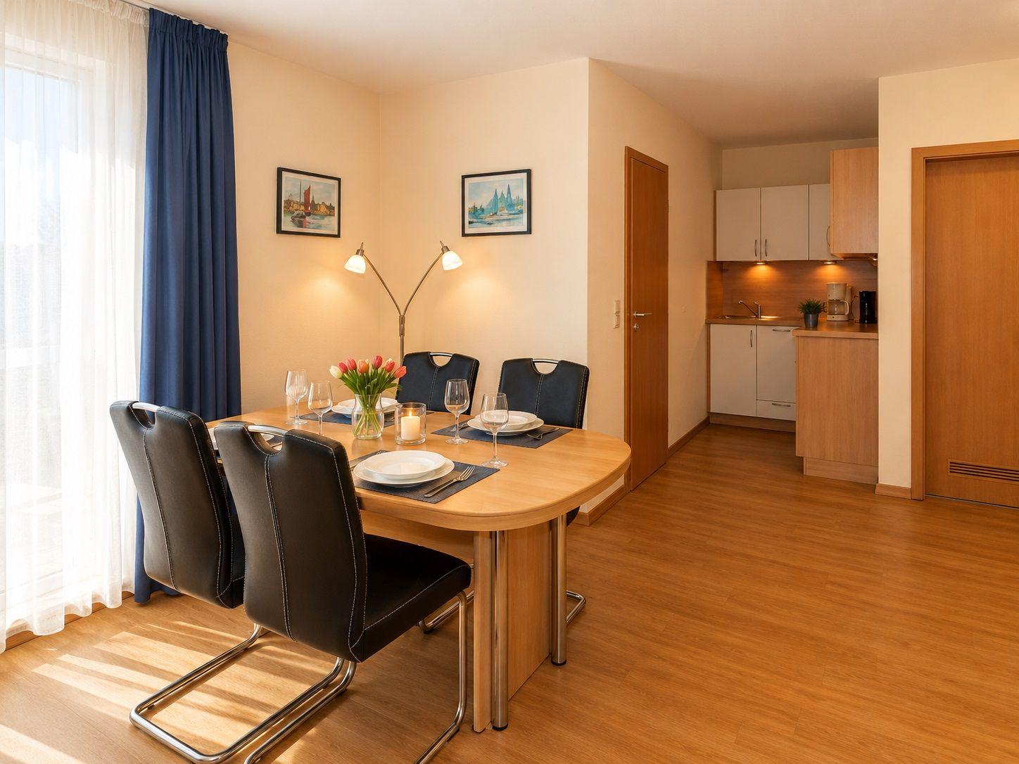 Dining area with table and chairs, kitchen in background, window with curtains.