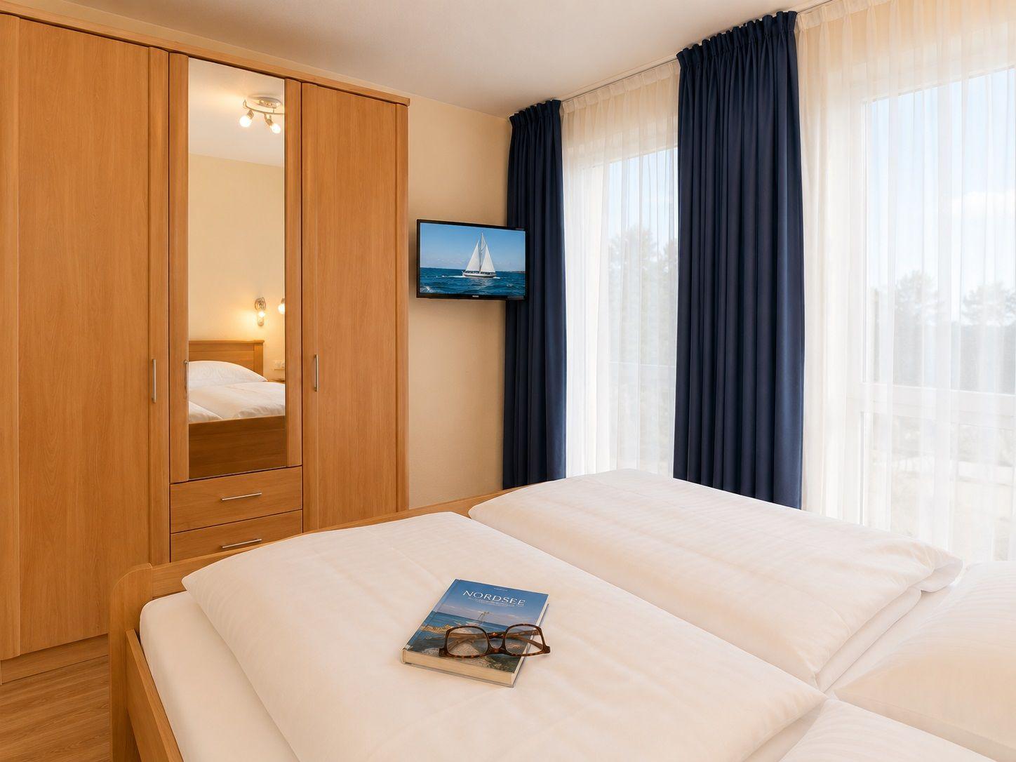 Double bed with book and glasses. Wooden wardrobe with mirror. TV on wall. Window with curtains.