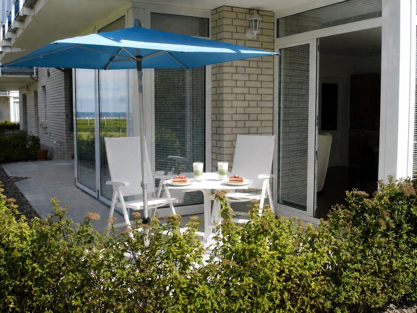 Terrasse mit blauem Sonnenschirm, weißen Stühlen und Tisch. Blick auf Grünfläche und Wasser.