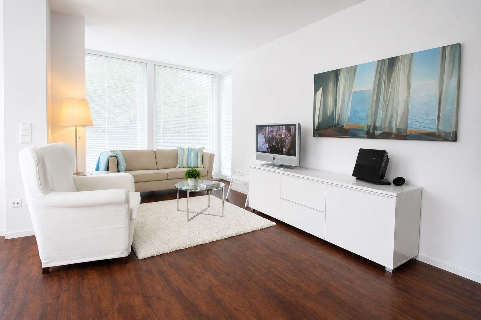 Wohnzimmer mit Sofa, Sessel, Fernseher und Wandbild. Holzboden und Fenster mit Jalousien.