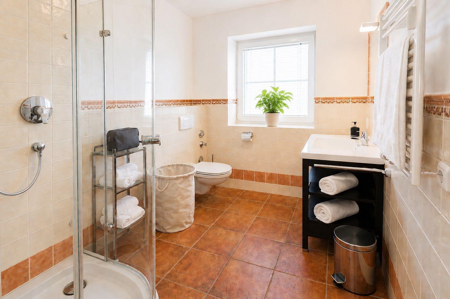 Badezimmer mit Dusche, Toilette und Waschbecken. Fenster, Pflanze und Handtücher sichtbar.