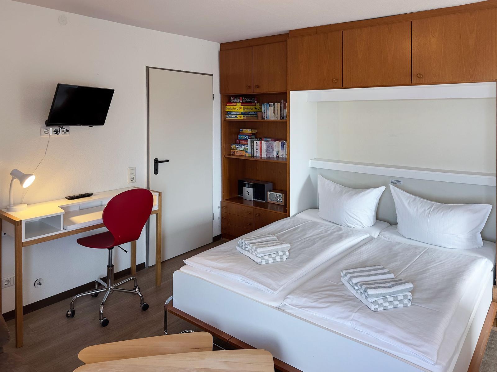 Zimmer mit Doppelbett, Schreibtisch, roter Stuhl und Wandfernseher.