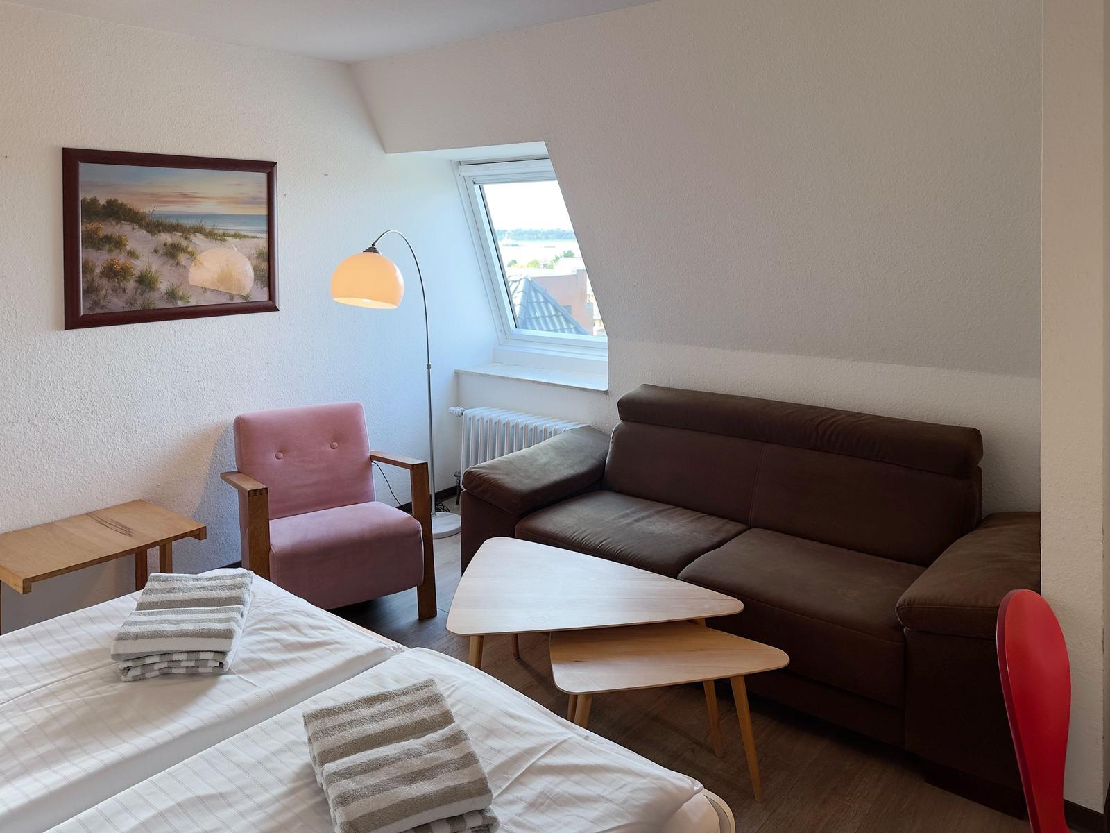 Zimmer mit Sofa, Sessel, Tisch und Bett. Fenster mit Blick auf Dach.
