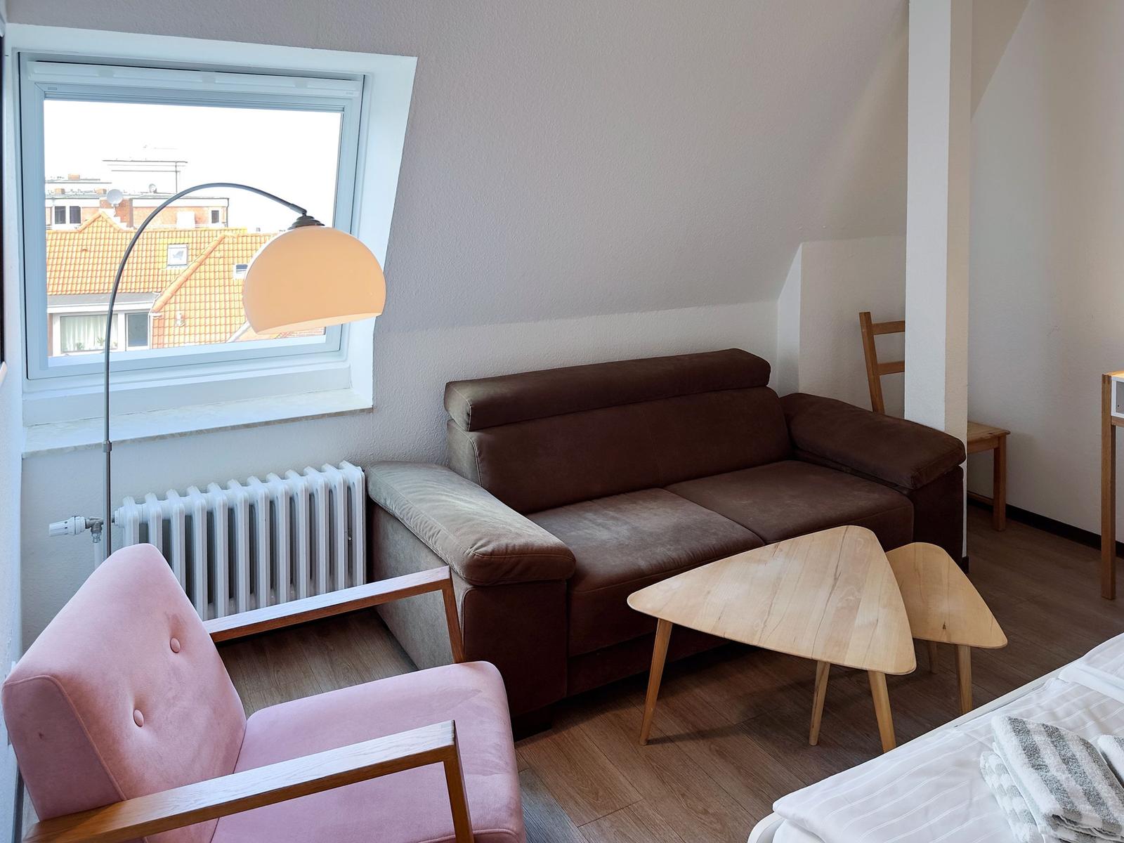 Wohnzimmer mit braunem Sofa, pinkem Sessel und Holztisch. Fenster mit Blick auf Dächer.
