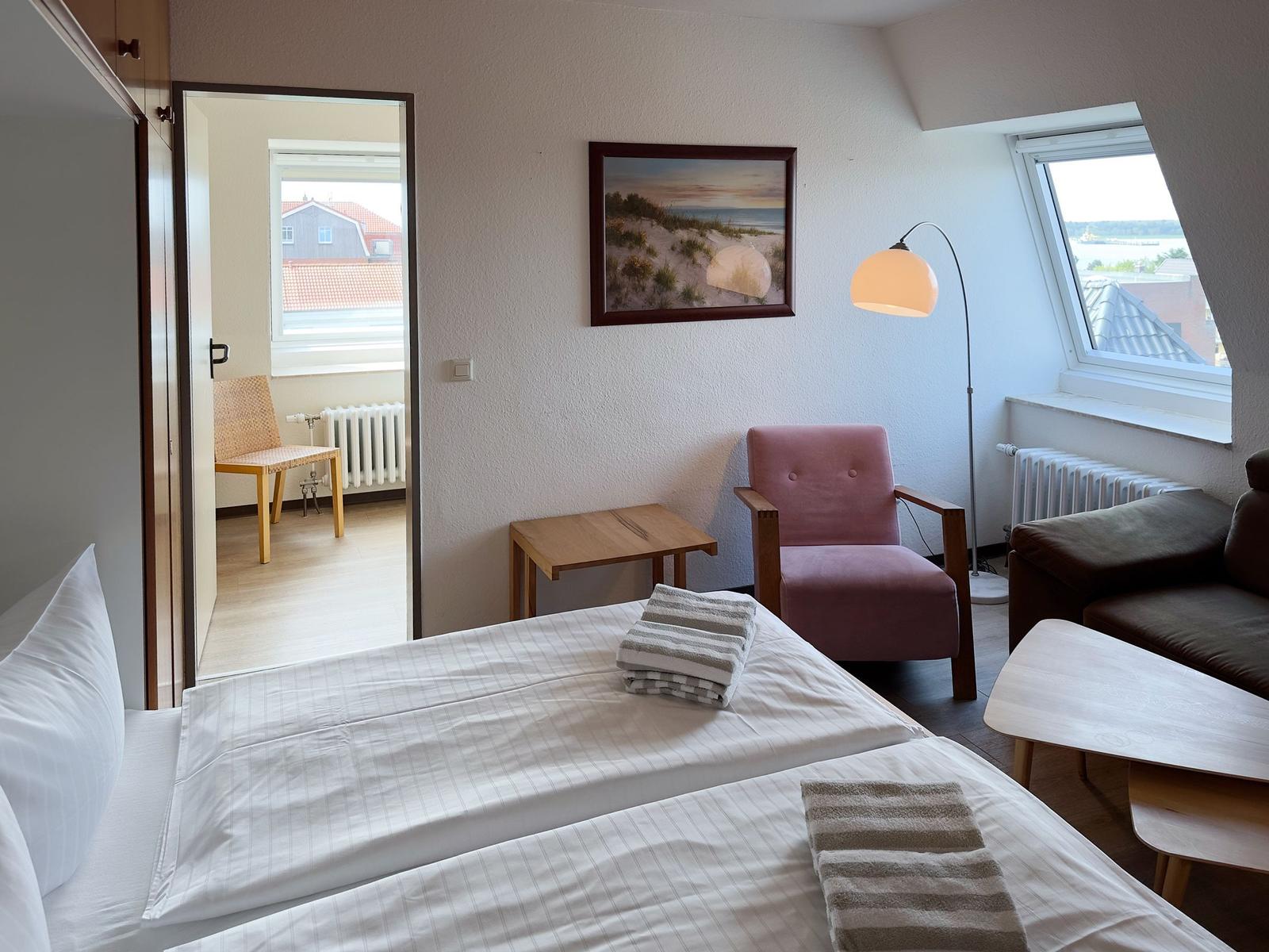 Zimmer mit Bett, Sessel und Fenster. Blick auf Dach und Wasser.