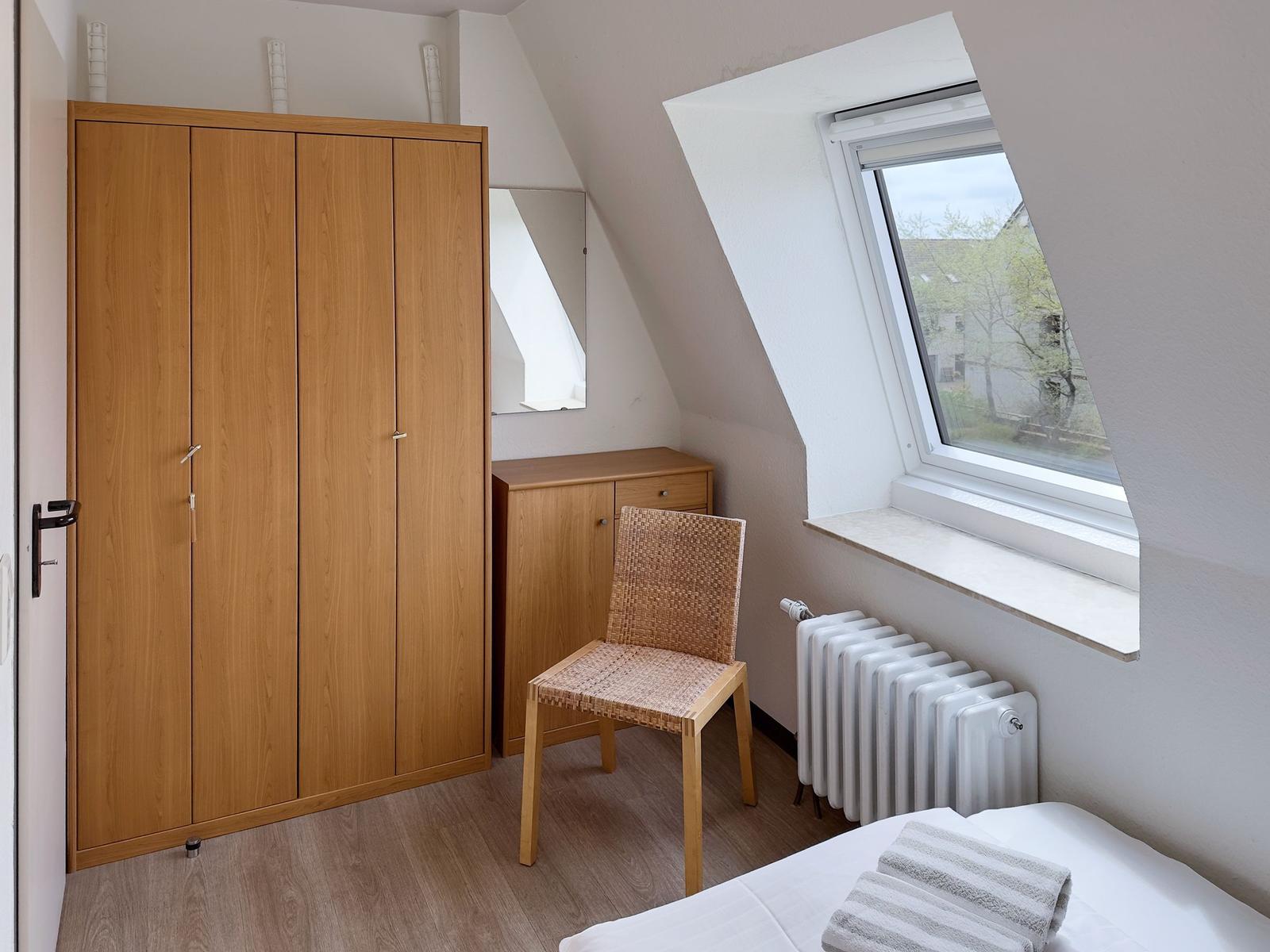 Schlafzimmer mit Dachschrägen, Kleiderschrank, Stuhl und Fenster.