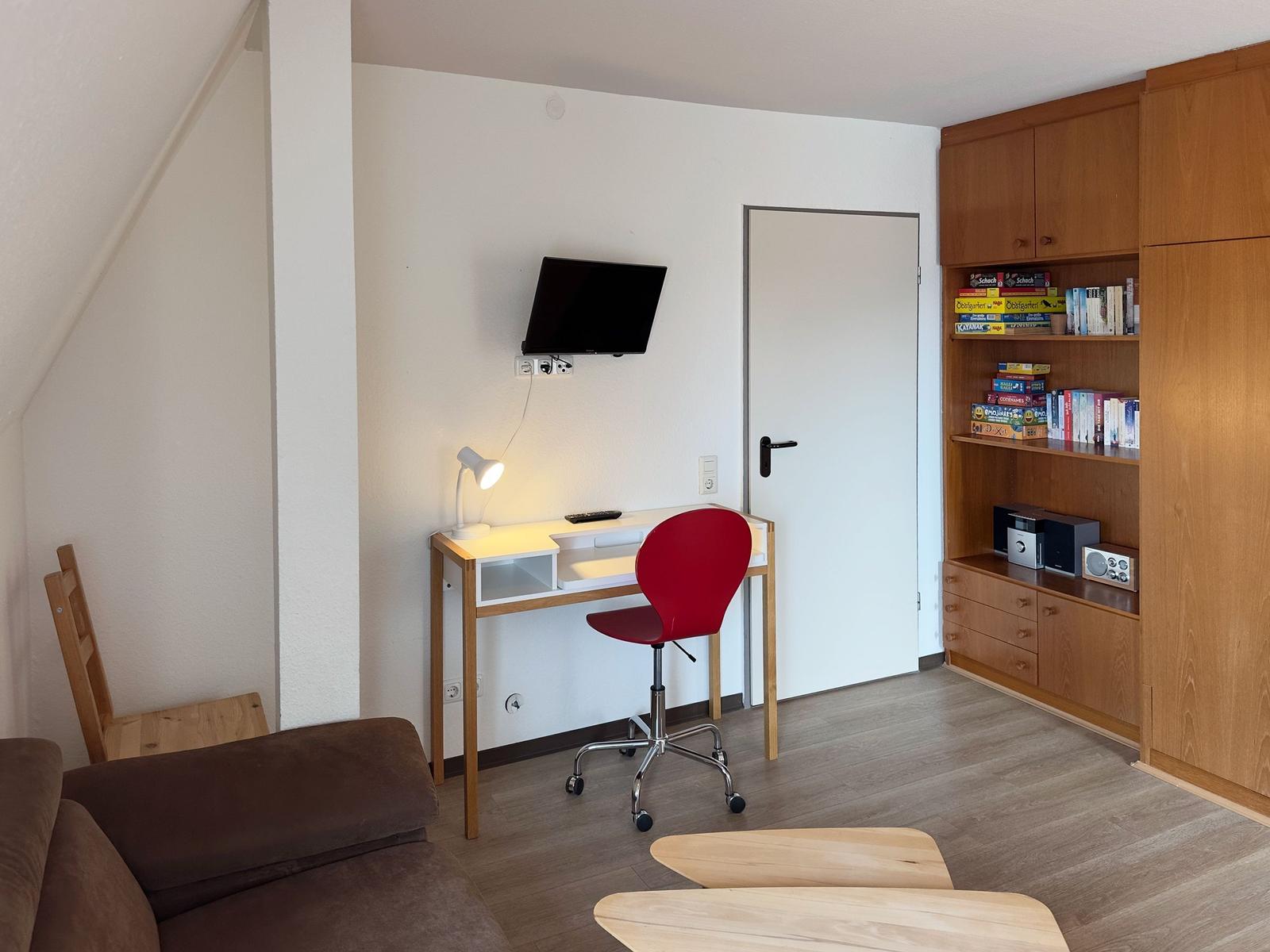 Schreibtisch mit roter Drehstuhl, Wandfernseher und Holzregal mit Büchern und Spielzeug.