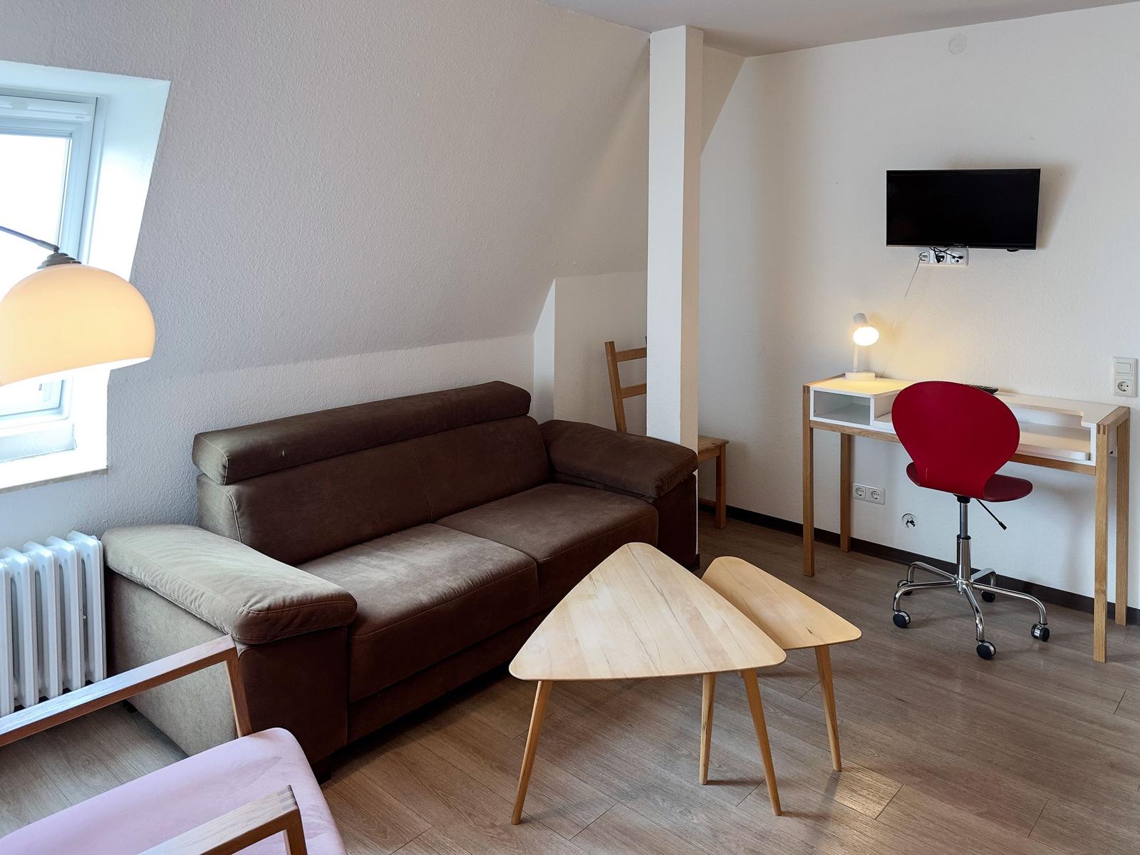 Ein Wohnraum mit braunem Sofa, Holztisch und roter Drehstuhl. An der Wand hängt ein Fernseher.
