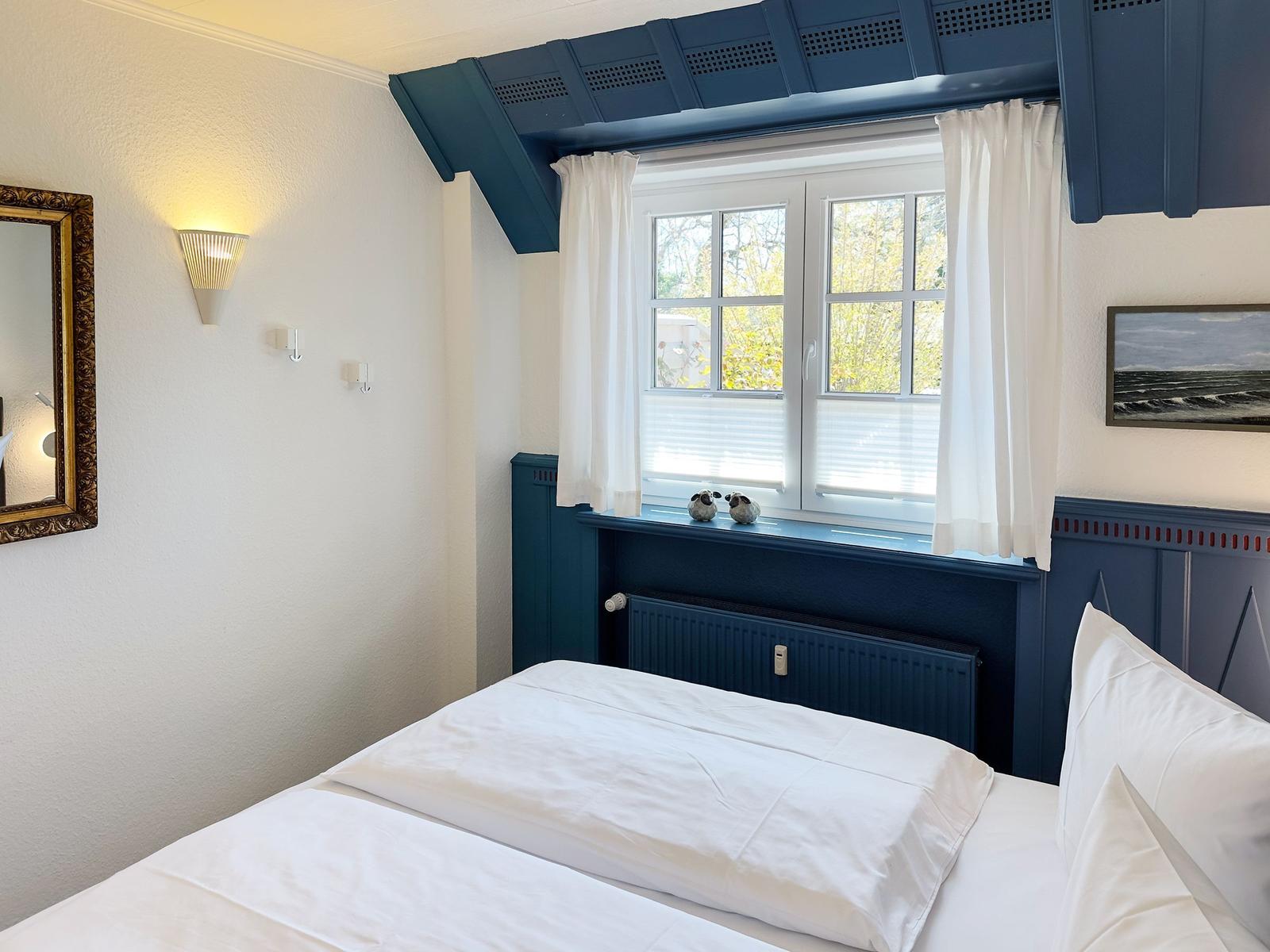 Ein Schlafzimmer mit weißem Bett, blauem Fensterbrett und Fenster mit weißen Vorhängen.
