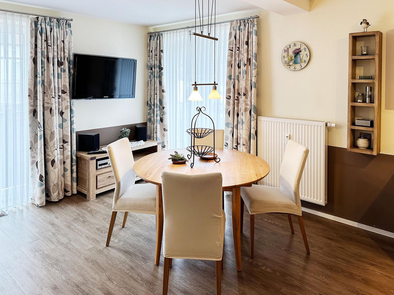 Rundes Esszimmer mit Holztisch, vier Stühlen und TV-Wand. Holzfußboden und Vorhänge.