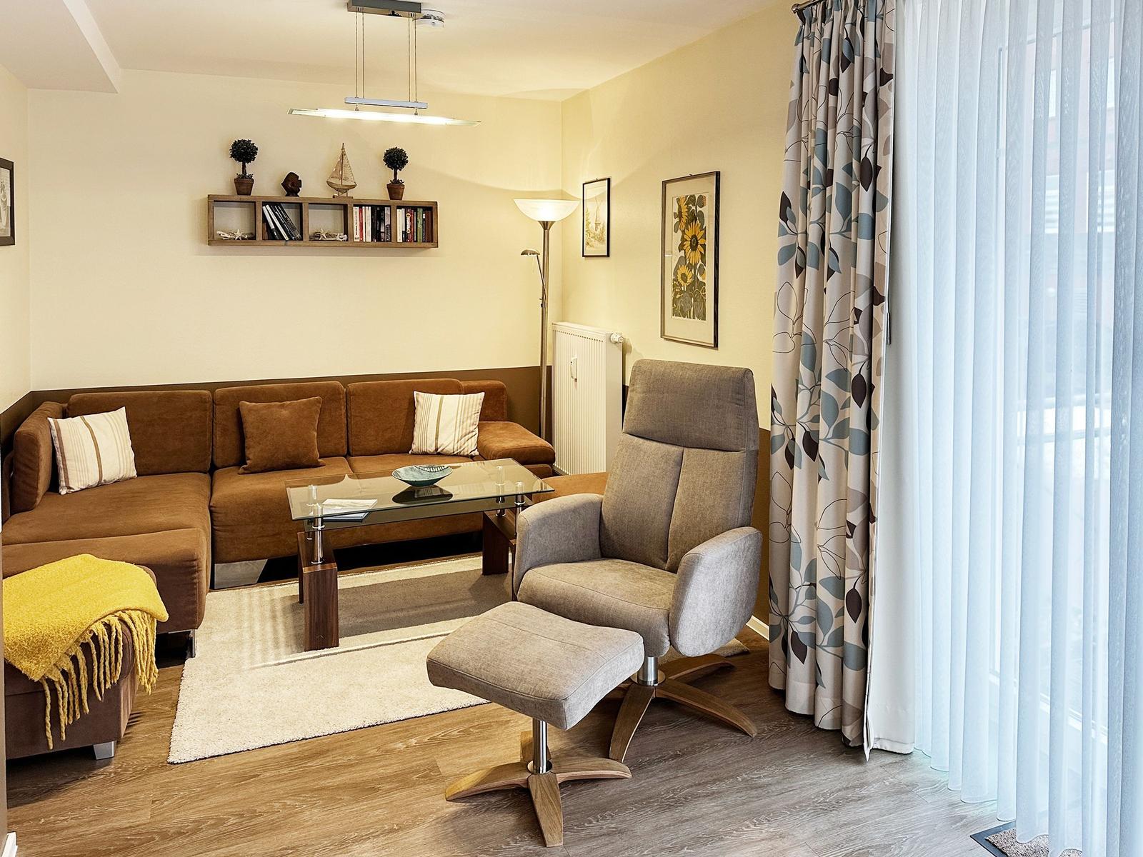 Ein Wohnzimmer mit braunem Sofa, grauer Sessel und Glas-Tisch. Vorhänge und Wandbild sind sichtbar.
