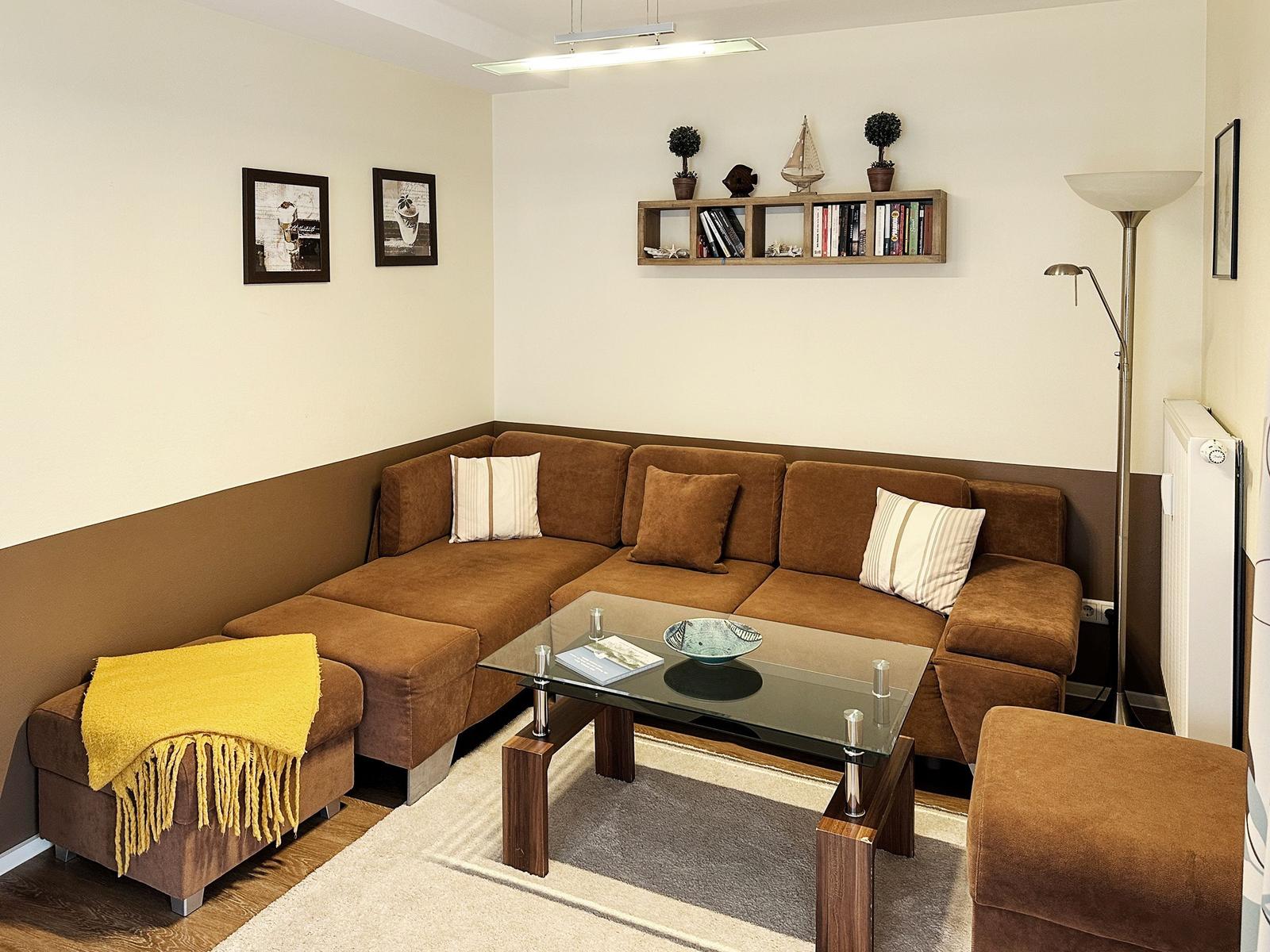 Ein Wohnzimmer mit braunem Sofa, Glas-Tisch und Wandregal mit Büchern.