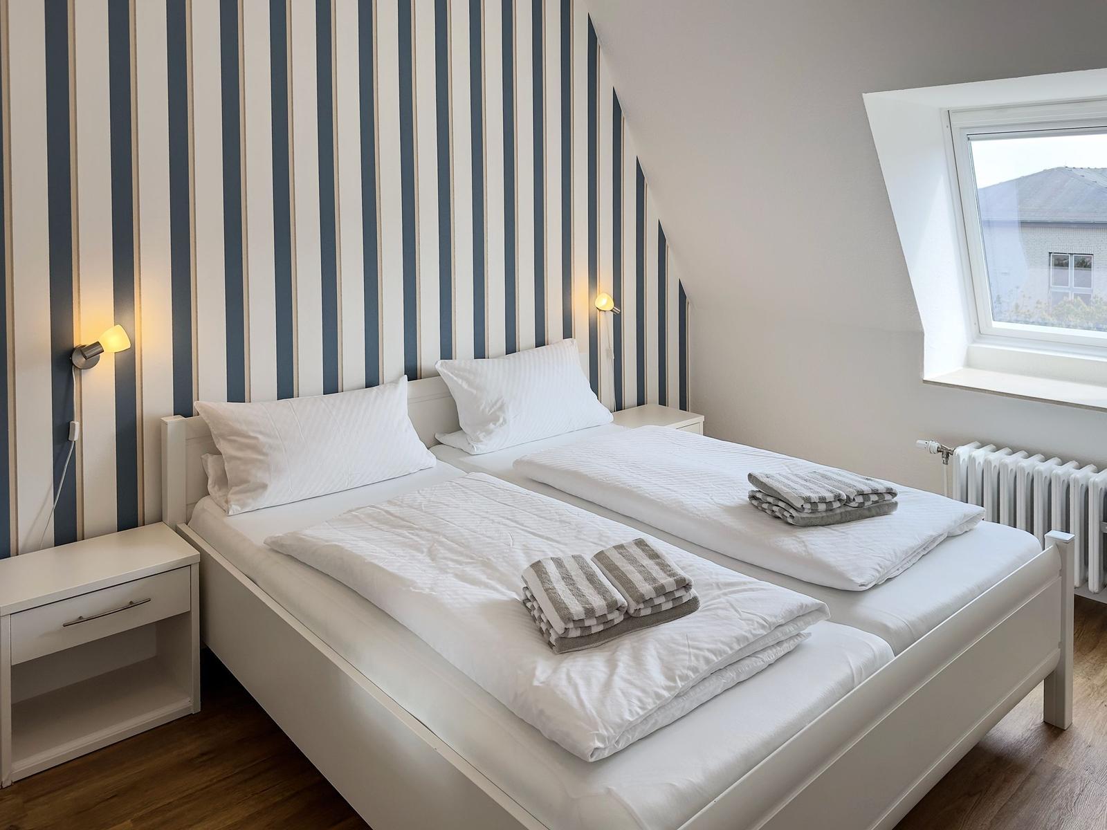 Two single beds with white linens and towels. Striped wall decor. Skylight.