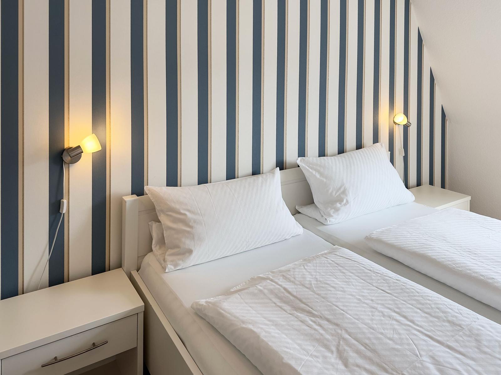 Two beds with white linens, striped wall, and nightstands with wall lamps.