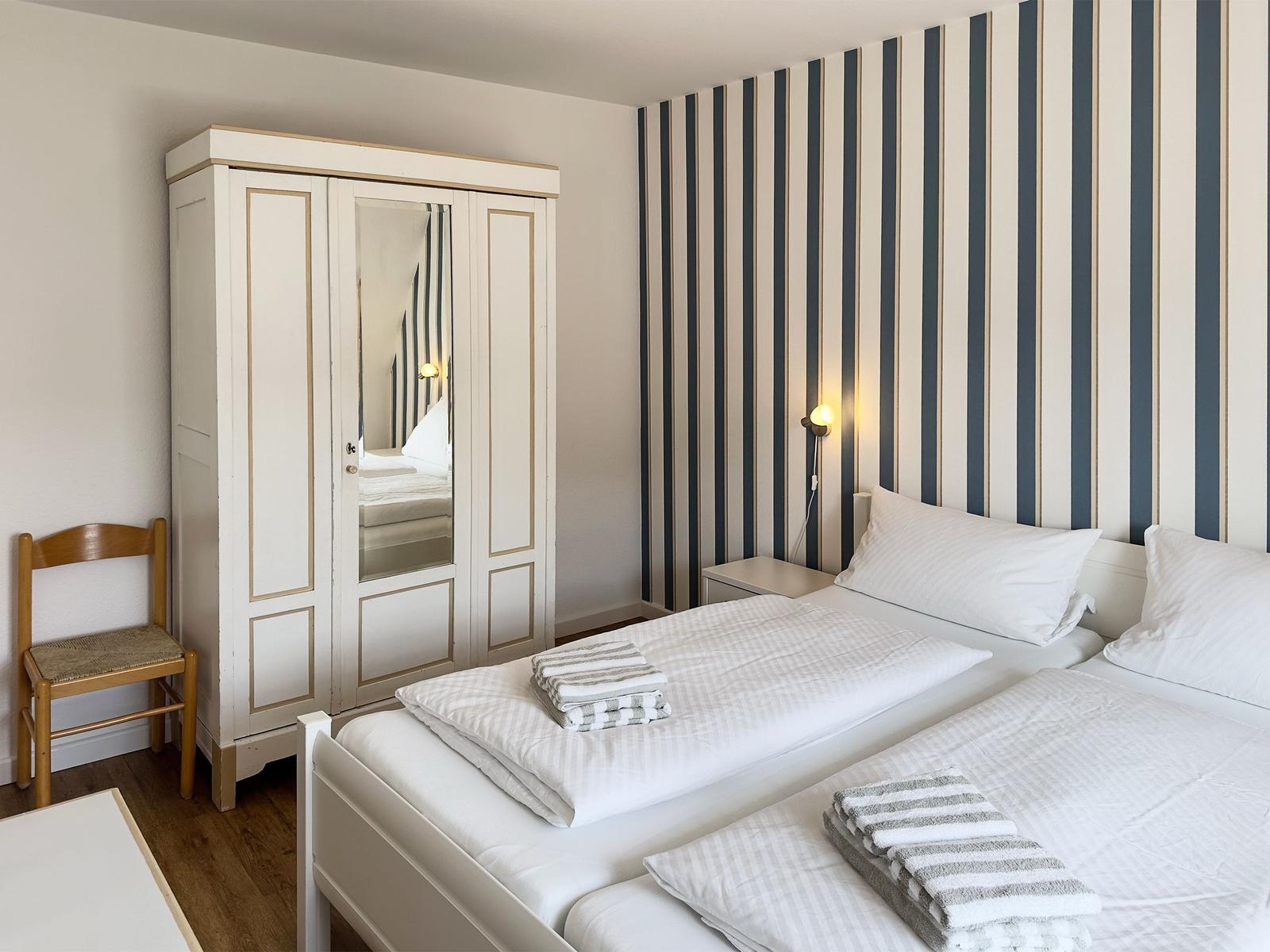 Two beds with white linens and striped towels. Striped wall and mirrored wardrobe.