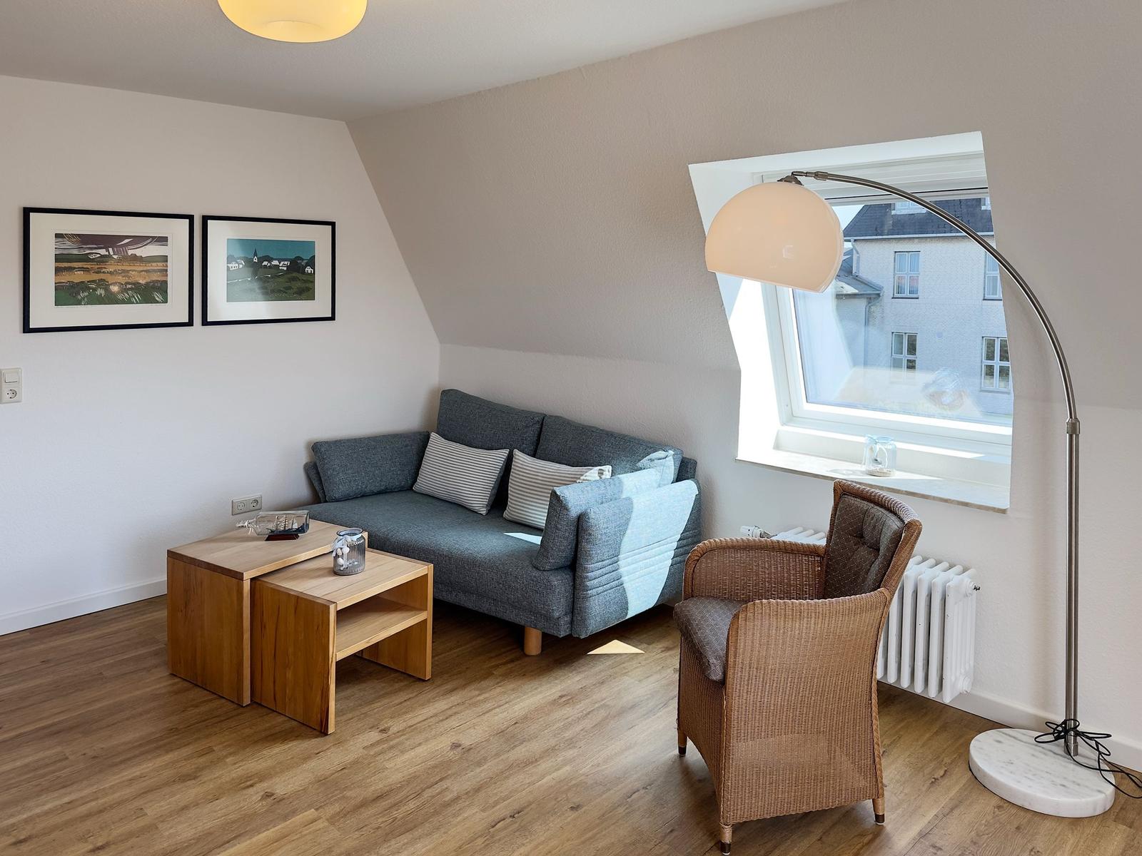 Living room with sofa, armchair, table, and window. Wall art and floor lamp are visible.