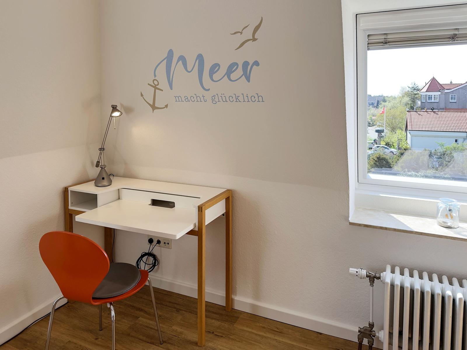 Desk with chair and wall decoration Meer macht glücklich. Window with view of residential area.