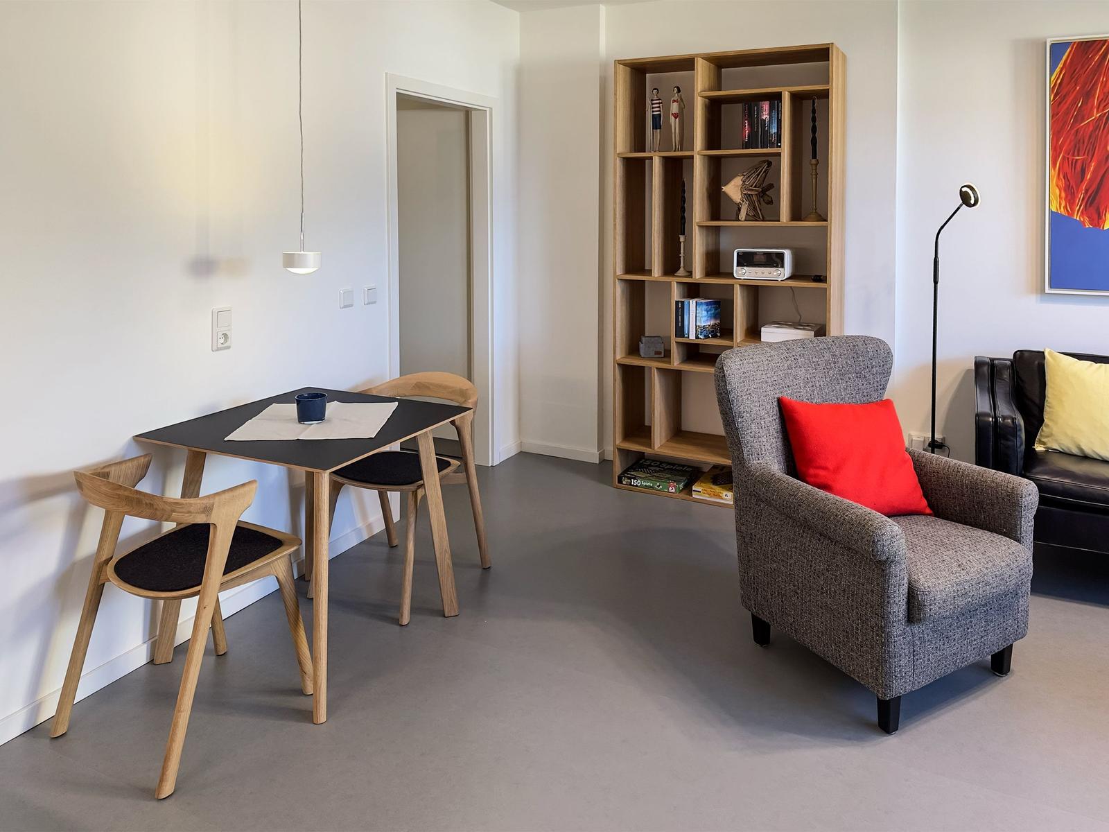 Dining area with table and chairs, armchair with pillow, bookshelf, and wall art.