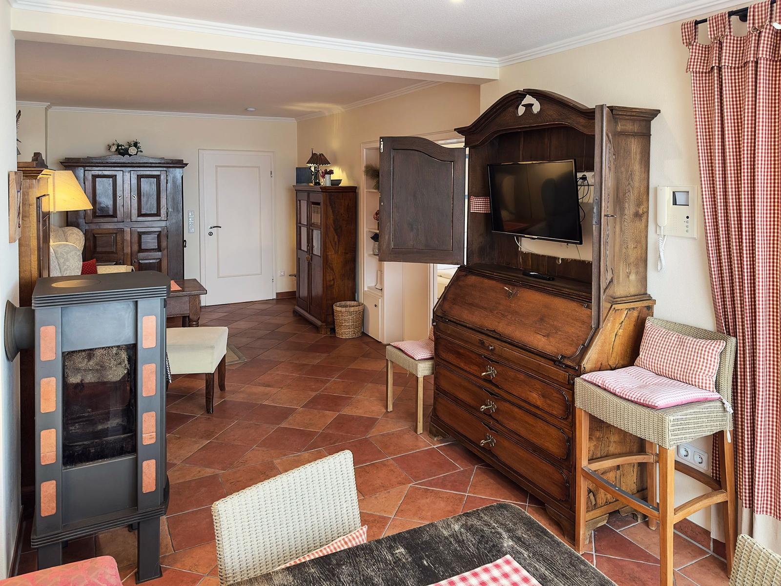 Wohnraum mit Holzmöbeln, Kaminofen und Fernseher in Schrank. Terrakottafußboden und Gittervorhänge.