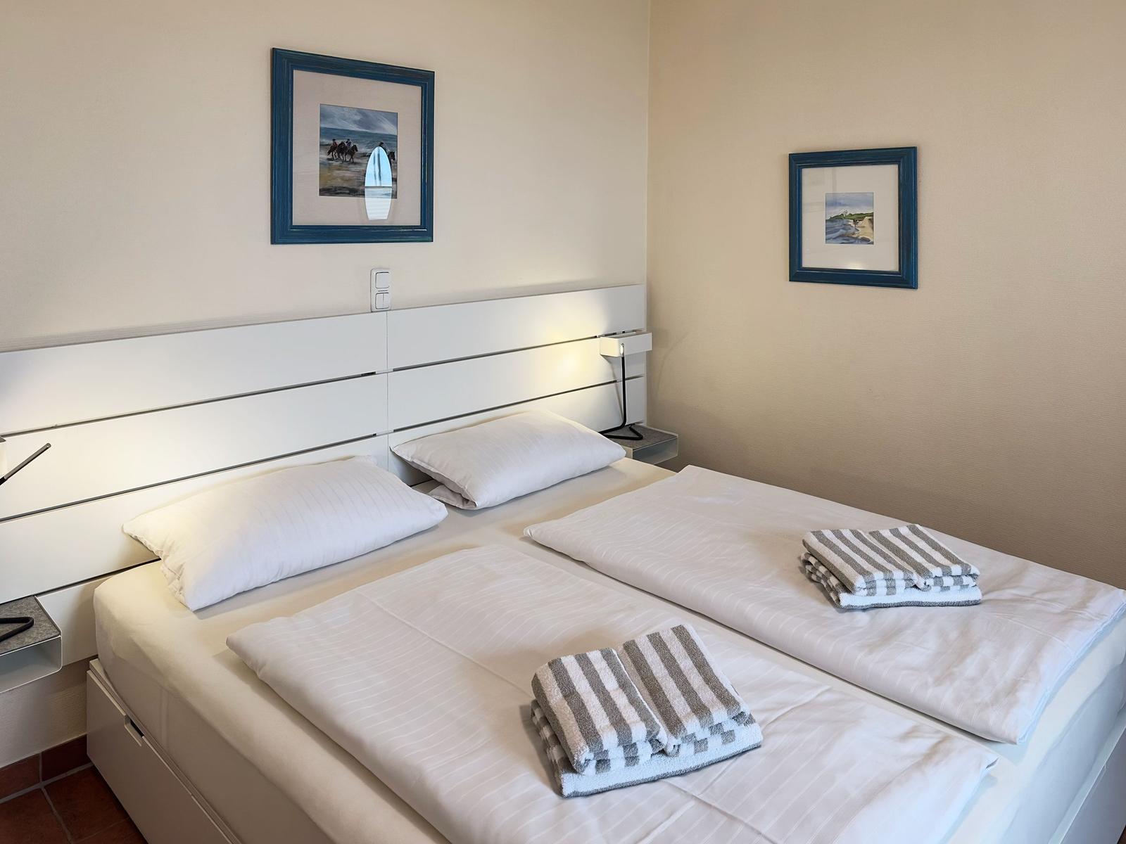Zimmer mit Doppelbett, weißen Bettwäsche und zwei Handtüchern. An den Wänden hängen Bilder.