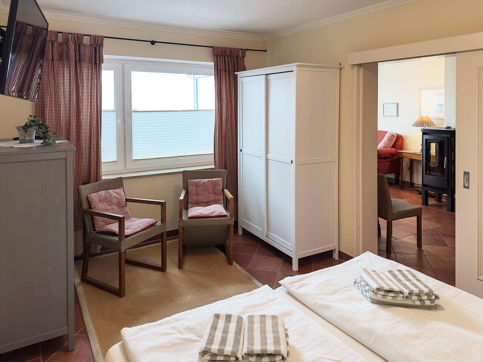 Zimmer mit Bett, zwei Stühlen, Schrank und Fenster. Durchgang zur Wohnfläche mit Kamin.