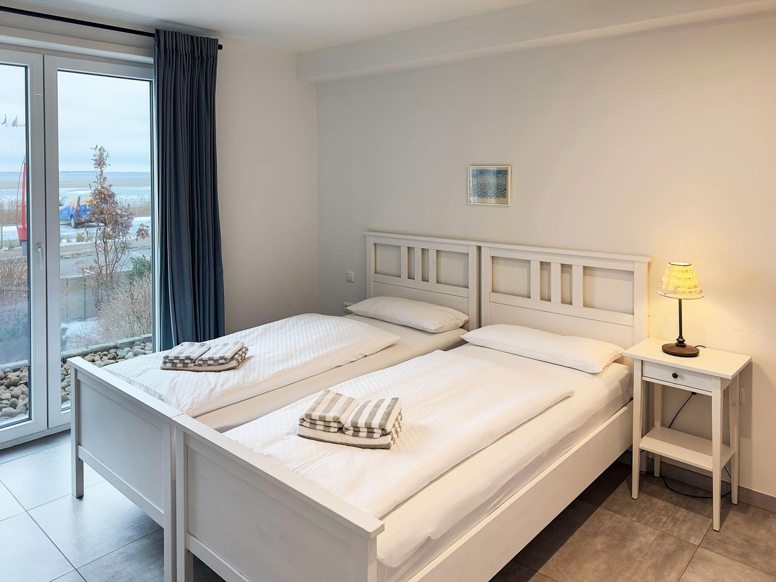 Zwei Einzelbetten mit weißen Laken und Handtüchern. Fensterblick auf Außengelände.