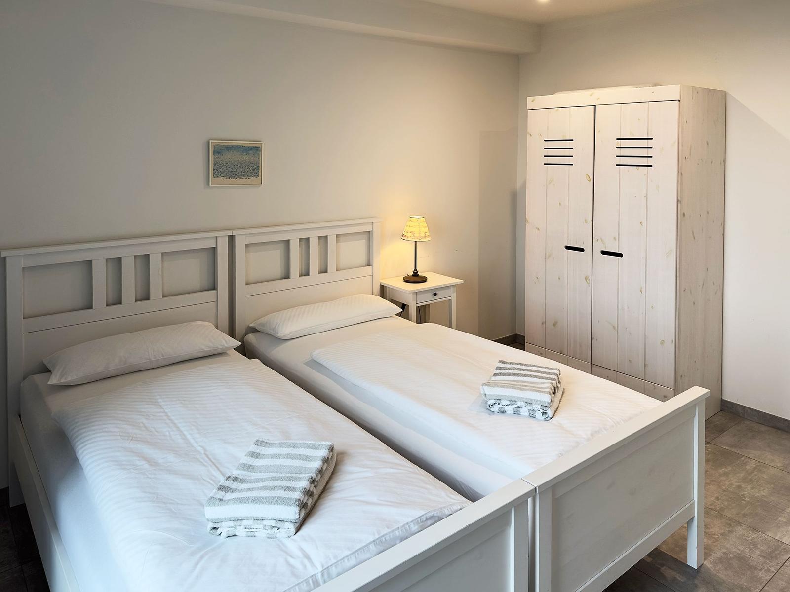 Zwei Betten mit weißen Bettwäschen, Nachttisch mit Lampe und Kleiderschrank.