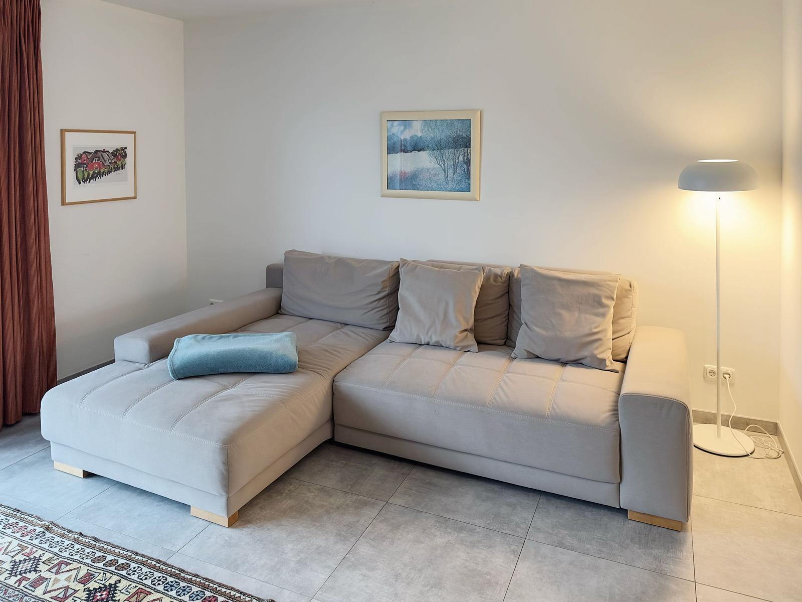 Ein Wohnzimmer mit grauem Ecksofa, zwei Bildern an der Wand und einer Stehlampe.