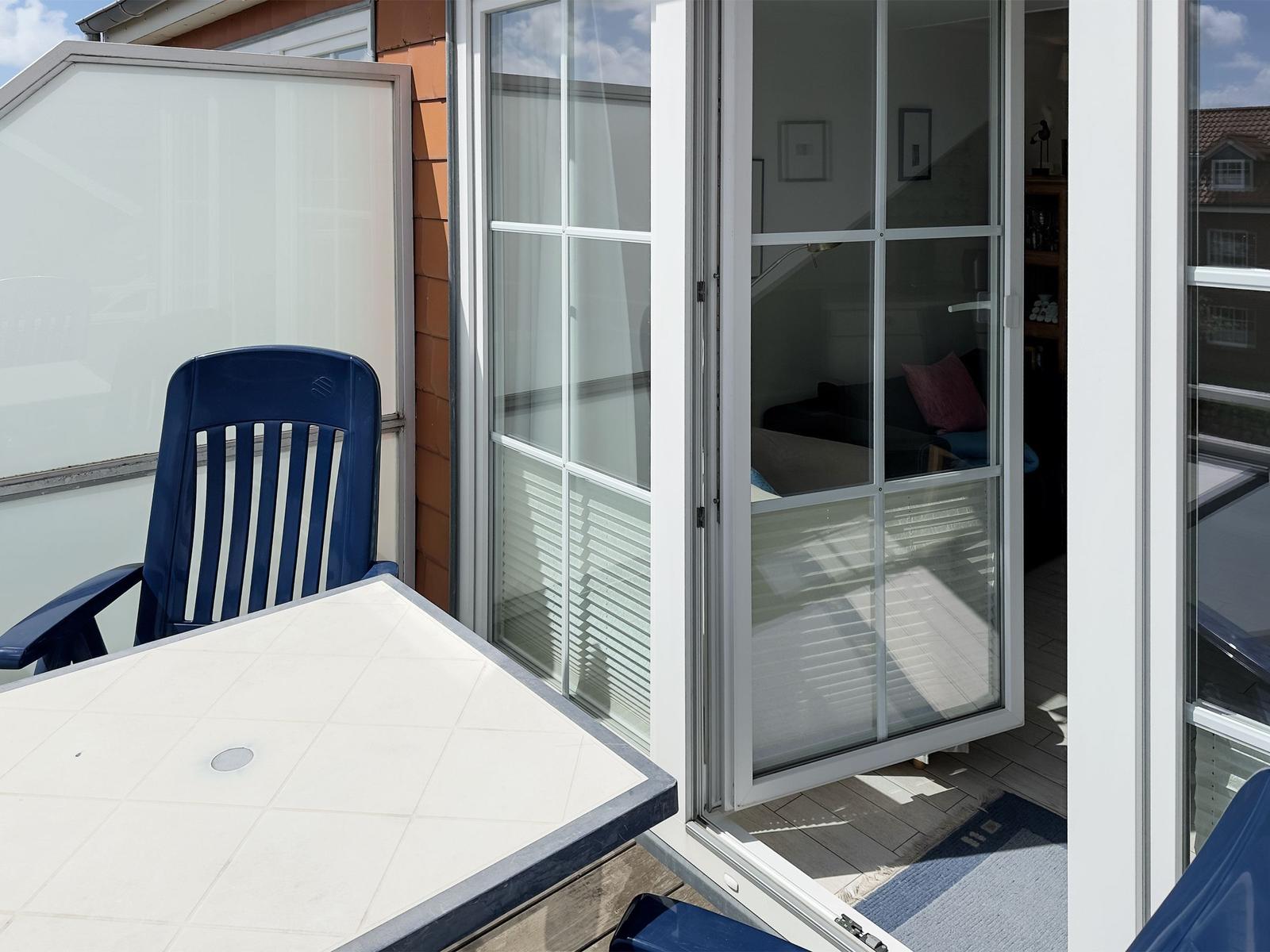 Terrasse mit Tisch und blauer Stuhl. Durchgehende Glastüren führen ins Wohnzimmer.