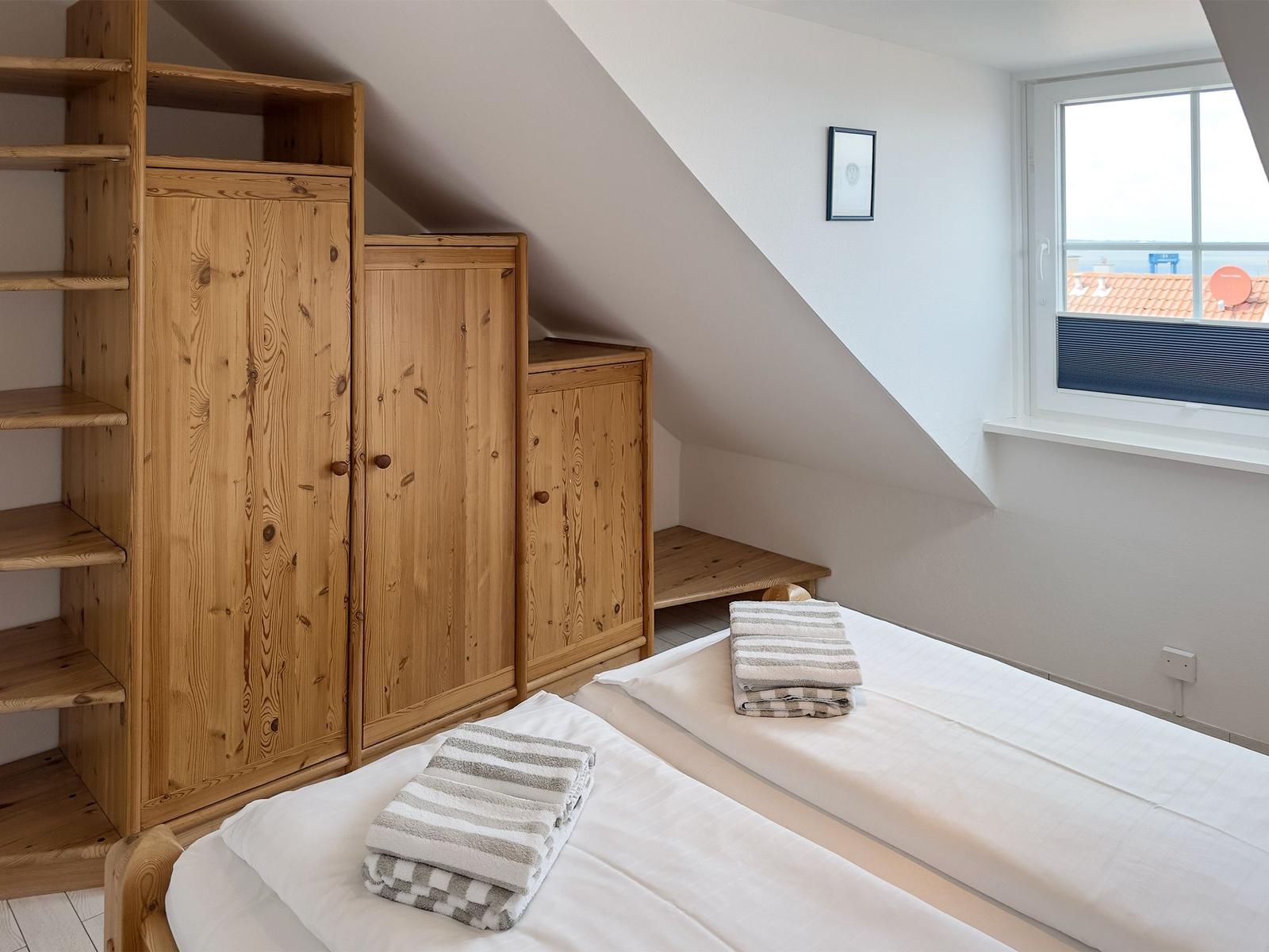 Zimmer mit Dachschrägen, Holzschrank und Bett mit Handtüchern. Fenster mit Blick auf Dächer.