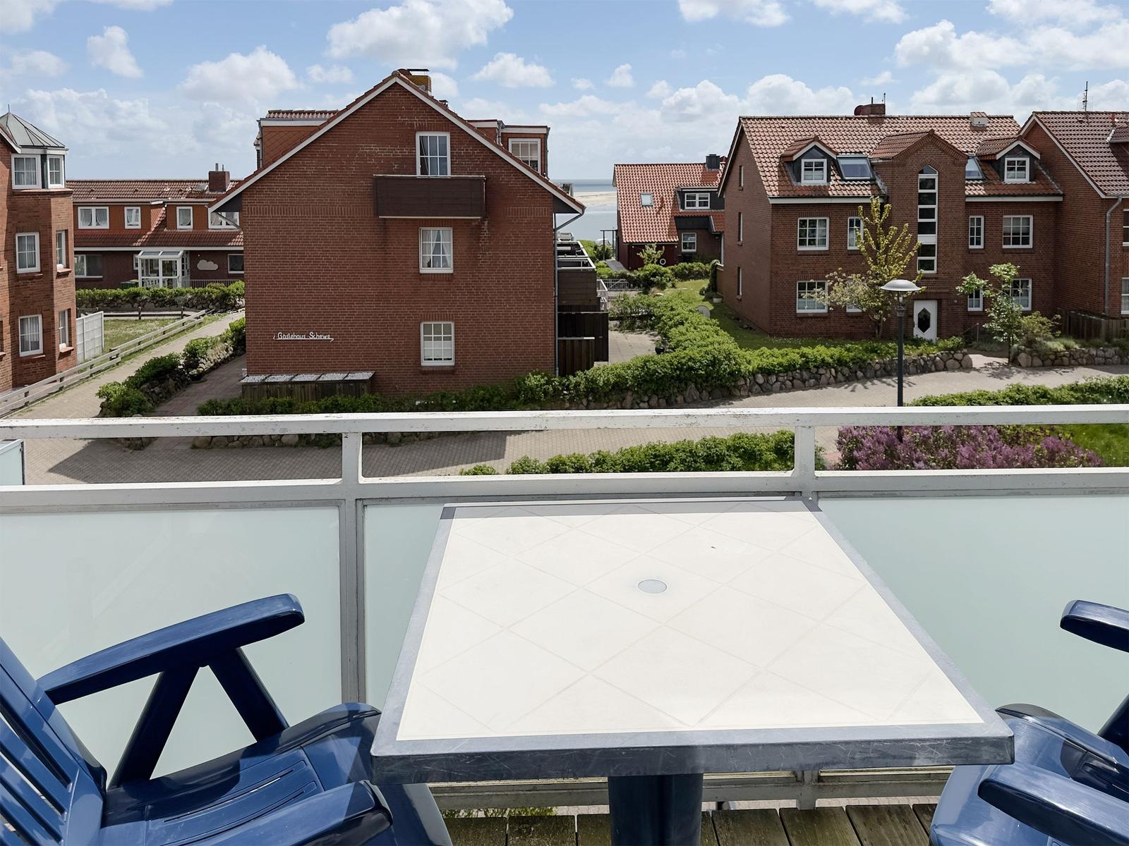 Terrasse mit Tisch und Stühlen. Blick auf rote Häuser und Strand.