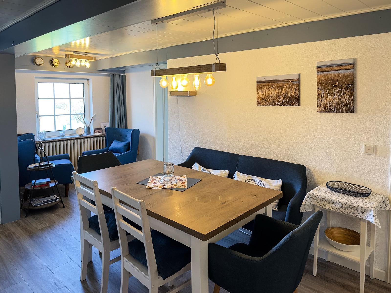 Essbereich mit Holztisch, Stühlen und Sofa. Wandbilder und Deckenleuchte sichtbar.