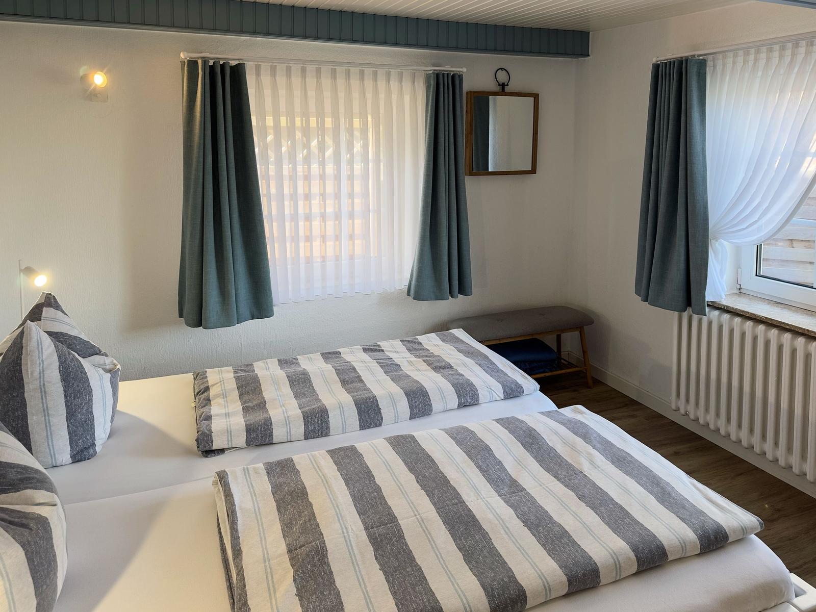 Zimmer mit zwei Betten, Fenstern mit Vorhängen und Spiegel an der Wand.