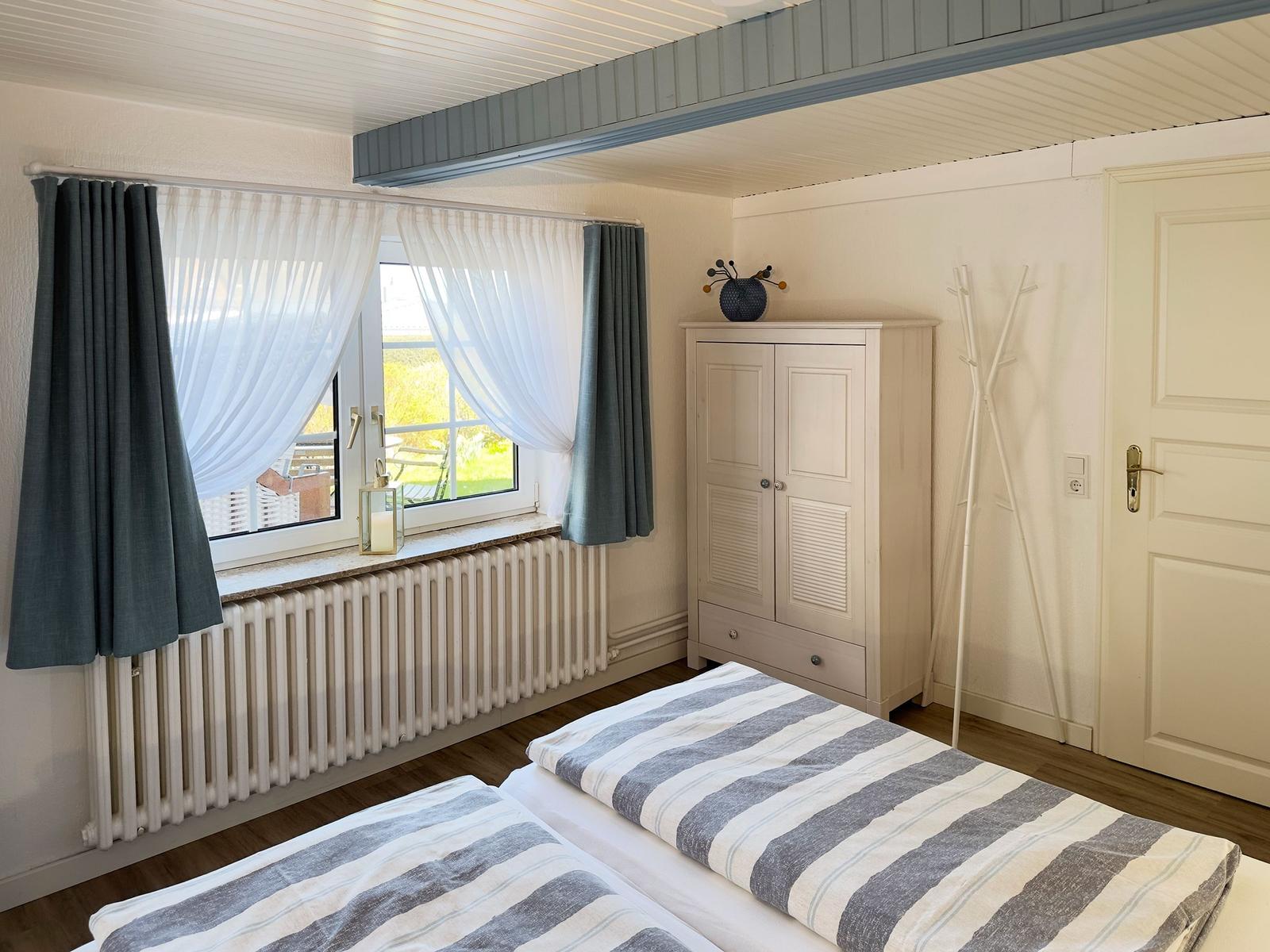 Zwei Betten mit gestreiften Bettwäsche, Fenster mit Vorhängen und Kleiderständer.