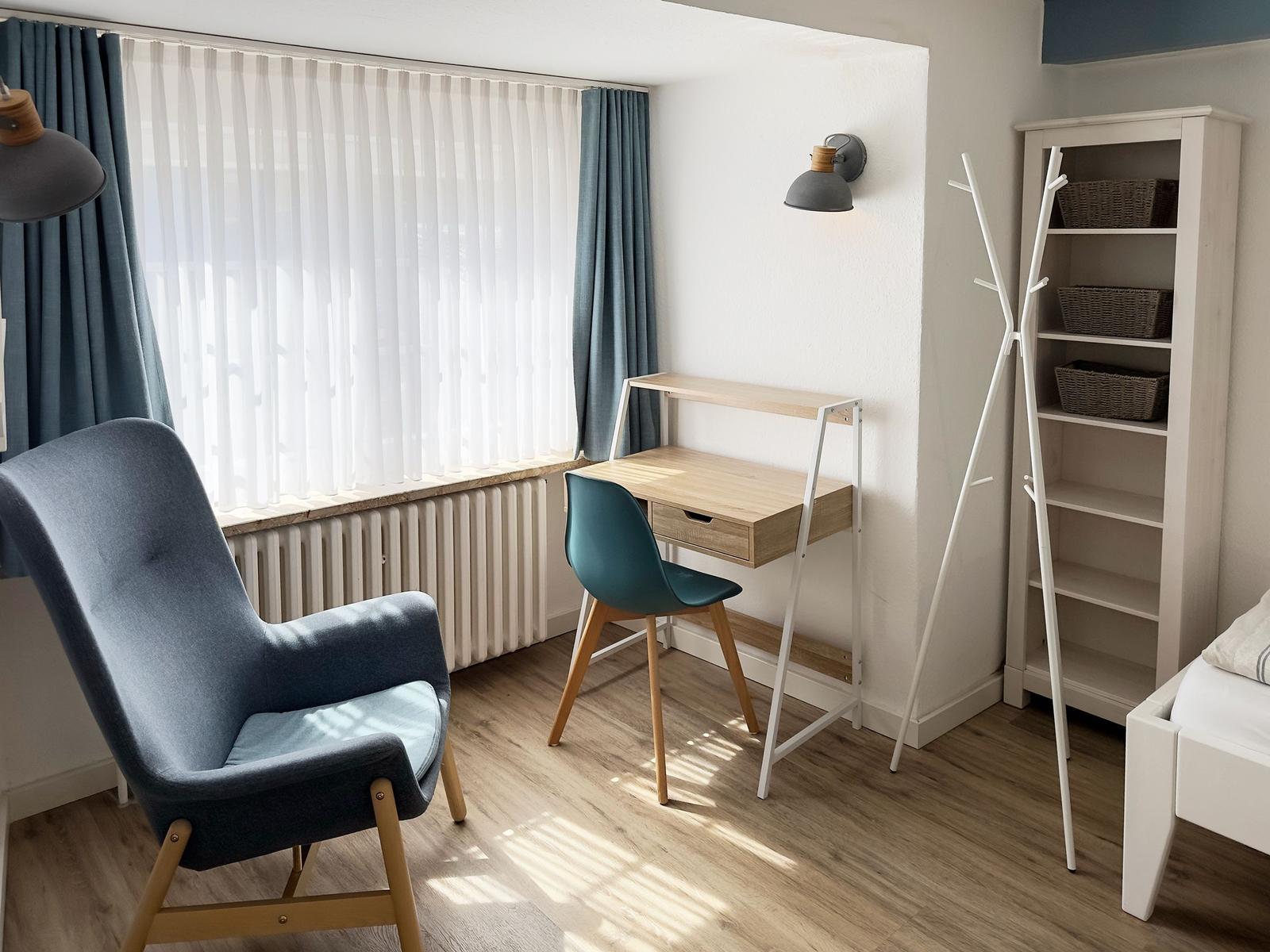 Ein Zimmer mit Schreibtisch, Stuhl, Kleiderständer und Fenster mit Vorhängen.